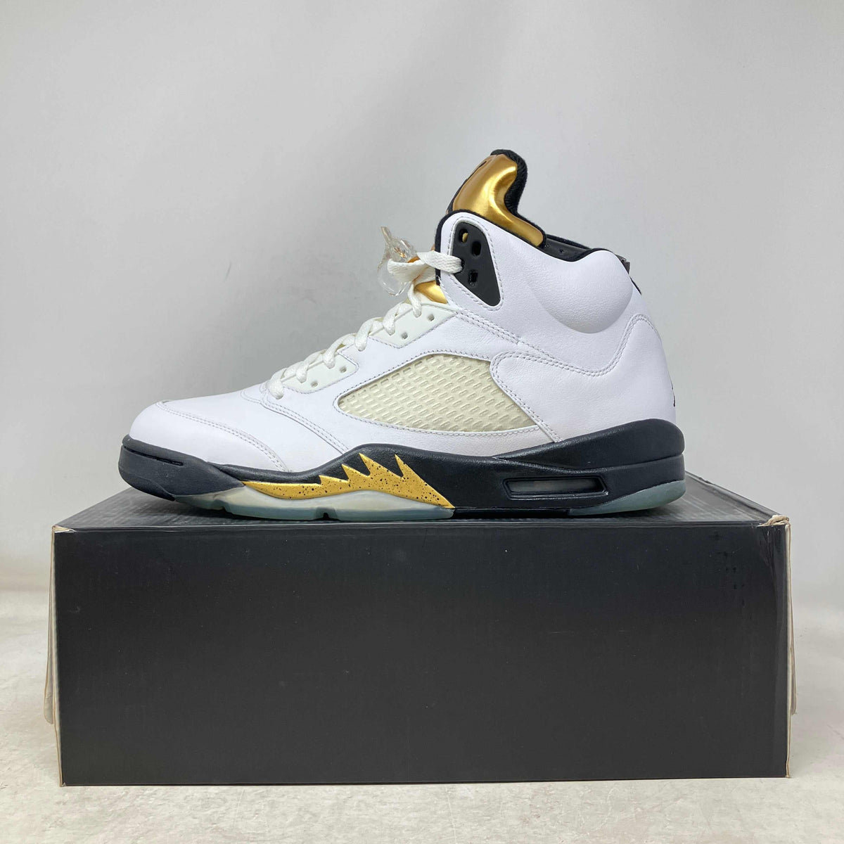 Jordan 5 Retro Olympic white leather sneaker with gold accents, black midsole, and mesh side panels on a black shoebox.