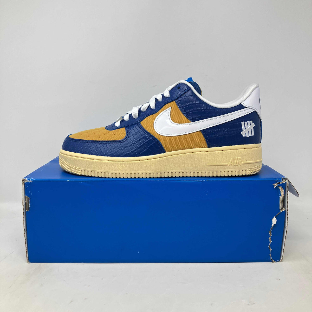 Nike Air Force 1 Low SP sneaker in blue and yellow croc-textured leather with white swoosh and logo accents on a beige sole.