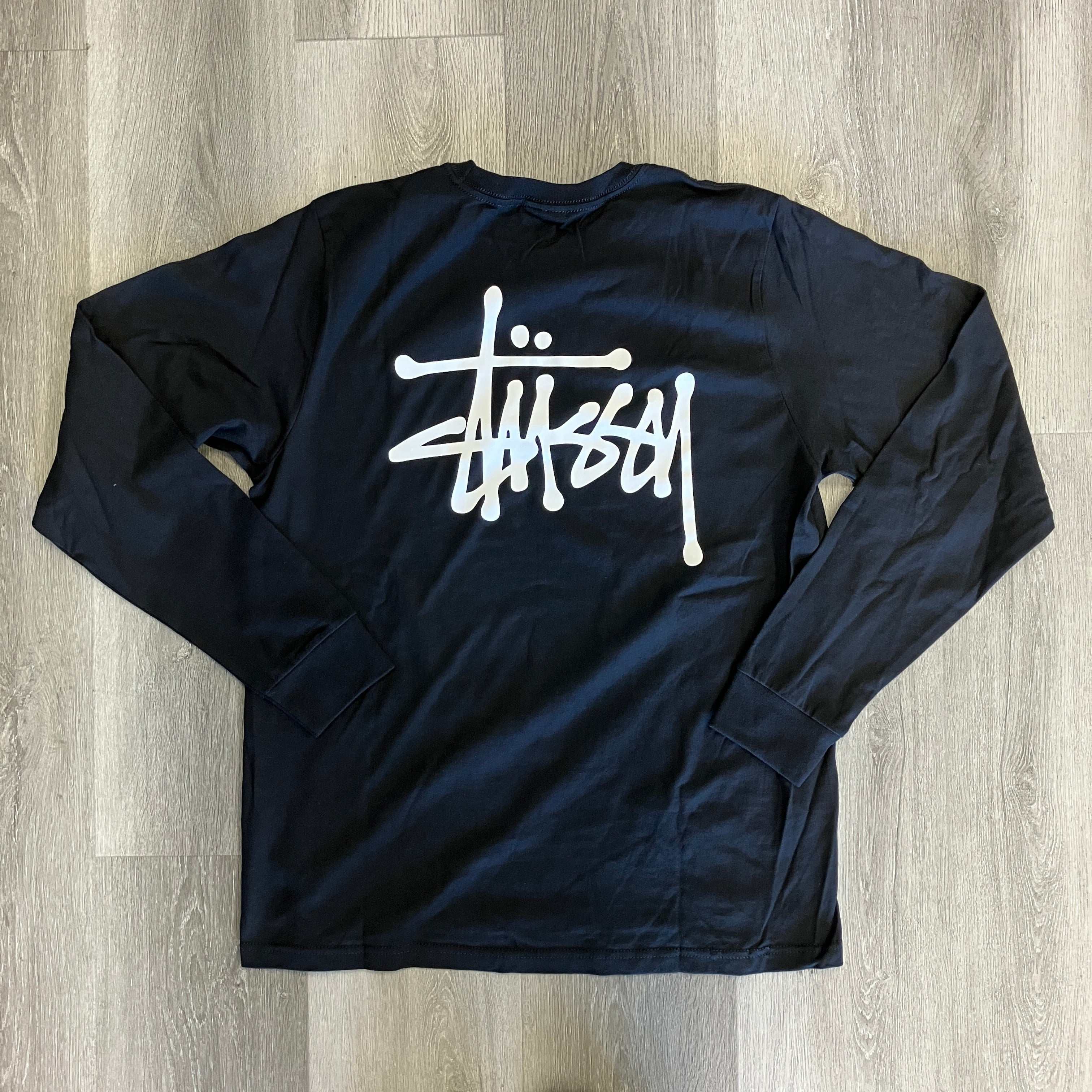 Black Stussy long sleeve tee with white logo on front, laid flat on wooden floor