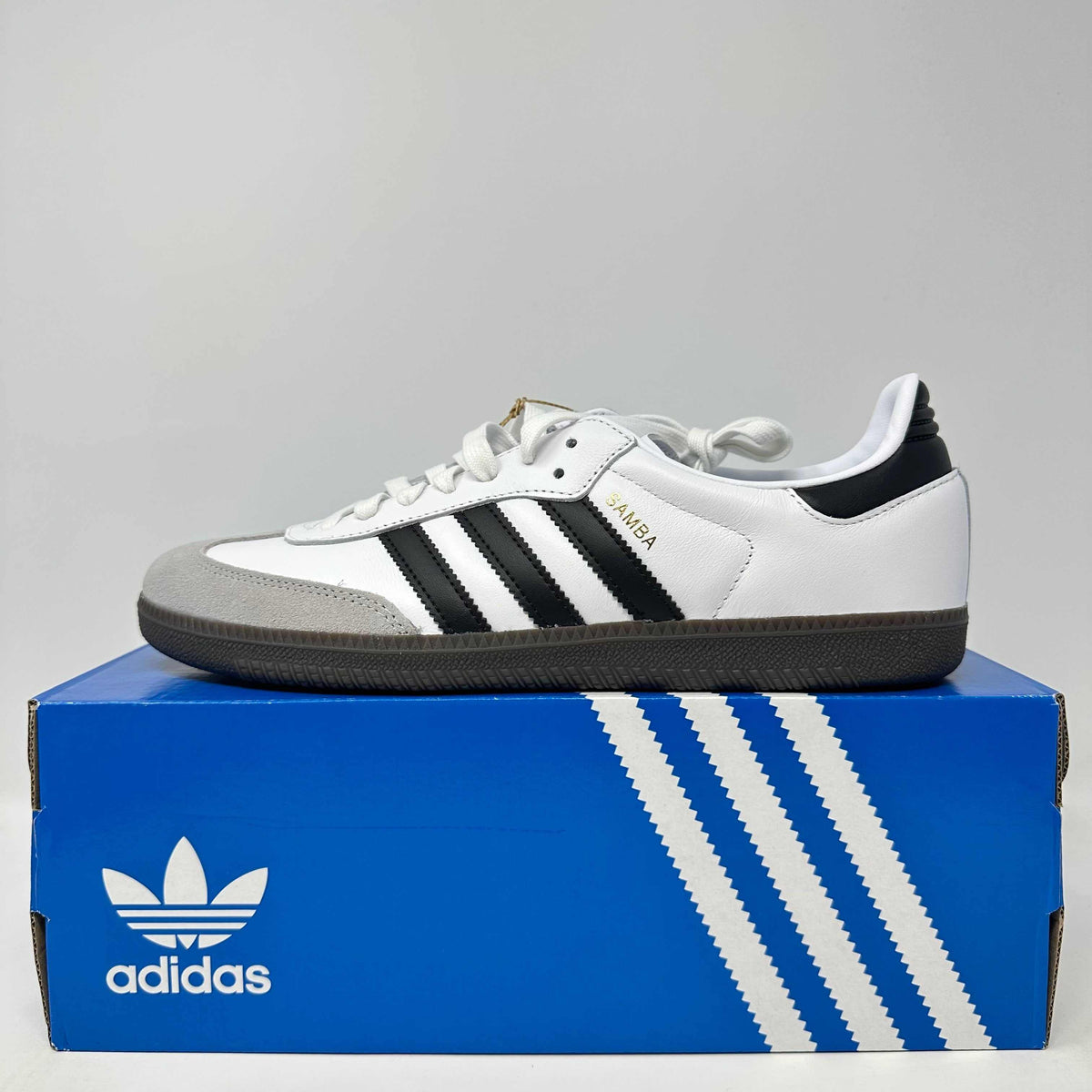 adidas Samba OG white leather sneaker with black stripes, gray suede toe, and gum sole on a blue adidas box