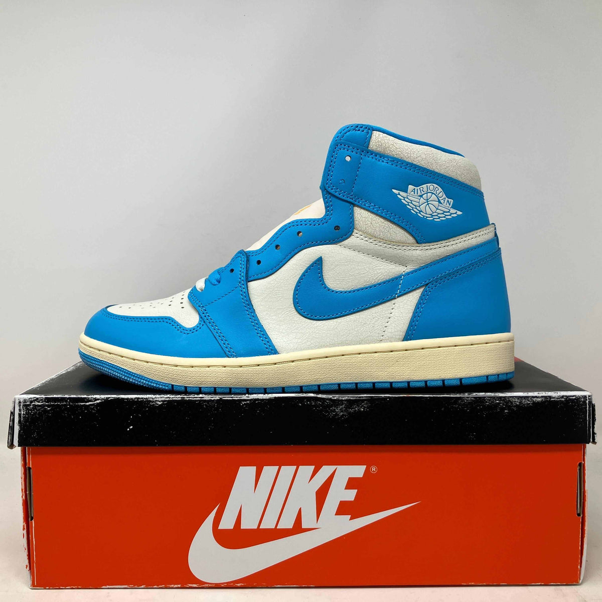 Jordan 1 Retro High OG sneaker in white and Carolina blue leather with vintage cream midsole on a black and red Nike box.
