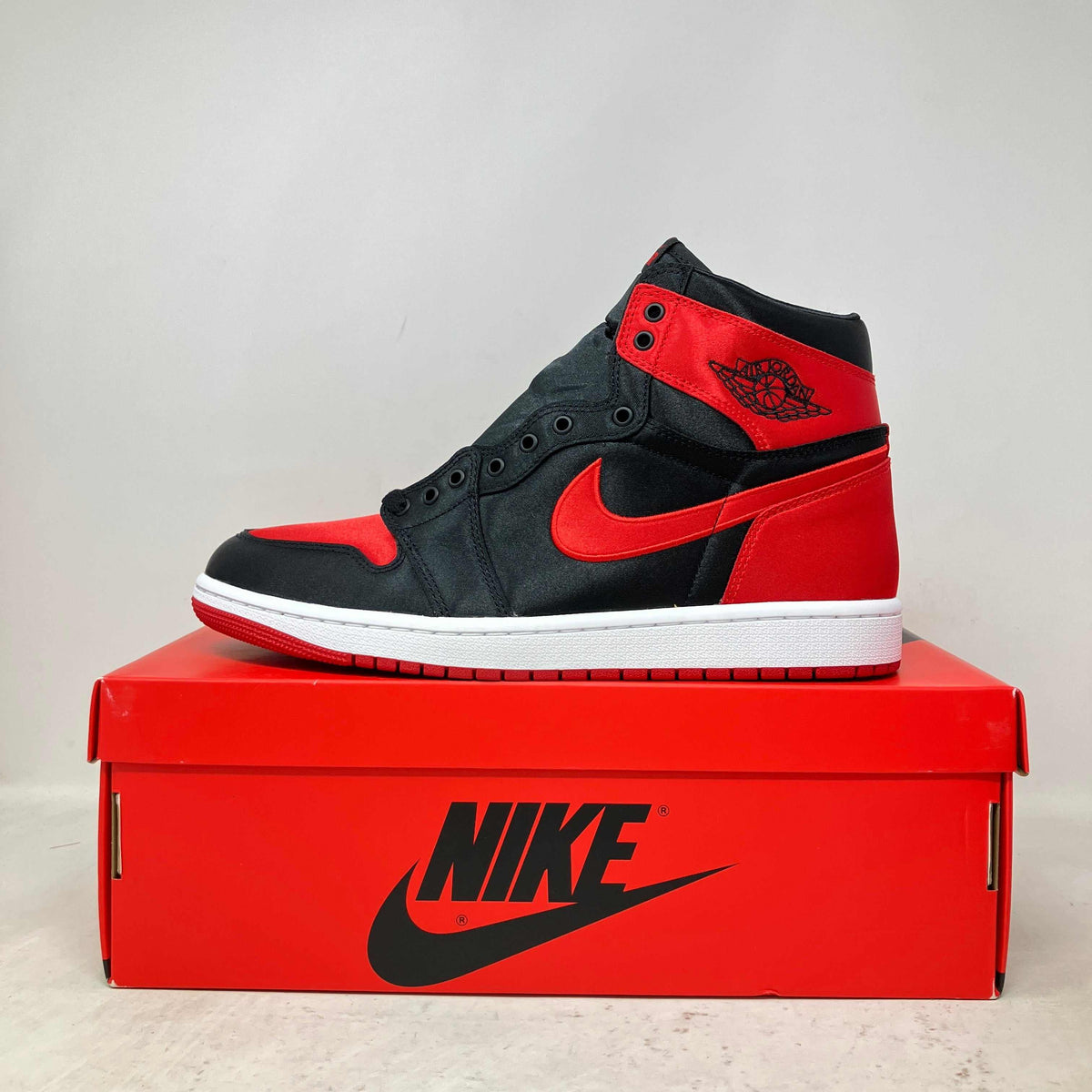 Jordan 1 Retro High OG sneaker in black and red satin material with a white sole and red Nike swoosh, displayed on a red Nike box.