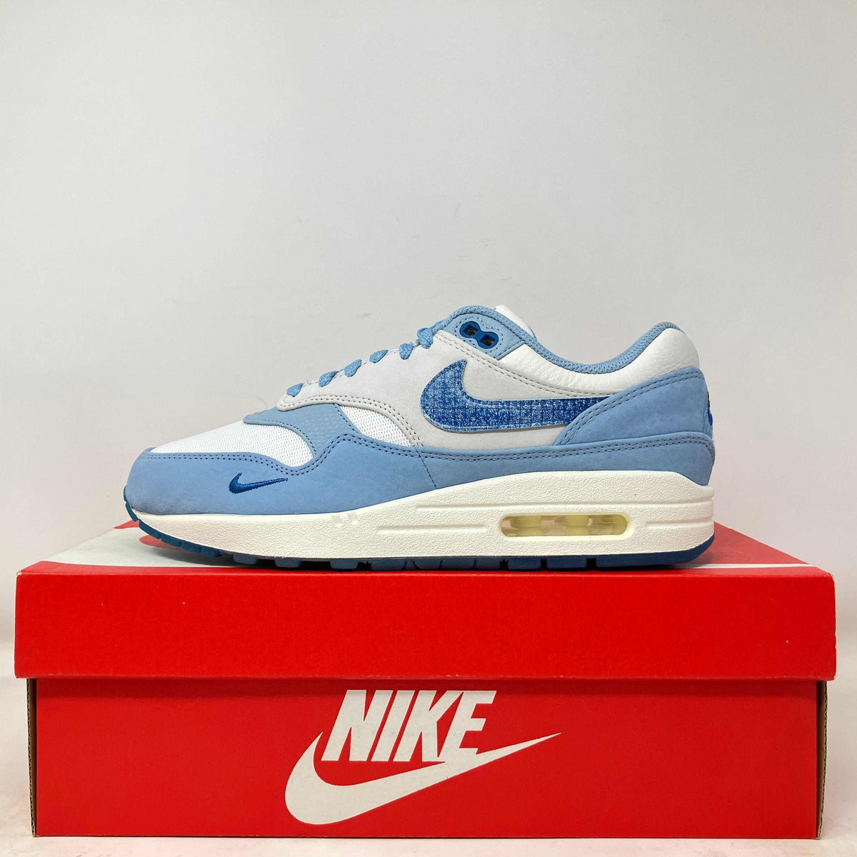 Nike Air Max 1 sneaker in white and light blue leather and mesh, with a textured blue Swoosh and visible Air unit.