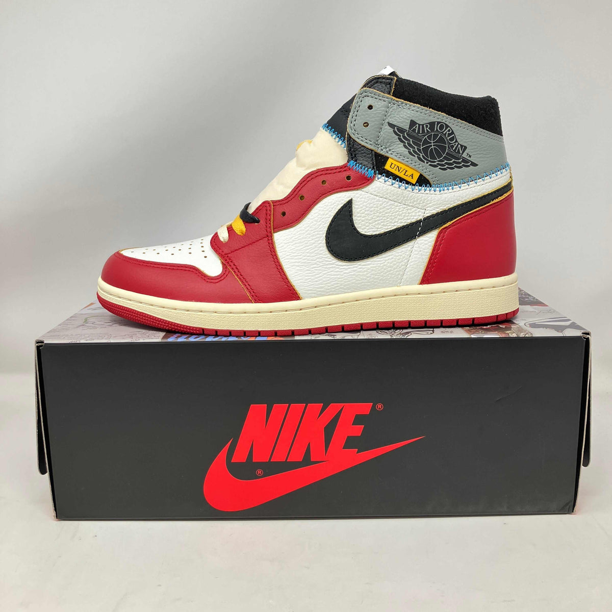 Jordan 1 Retro High OG sneaker in red, white, black, and grey leather with yellow and blue stitching details on a black Nike box.
