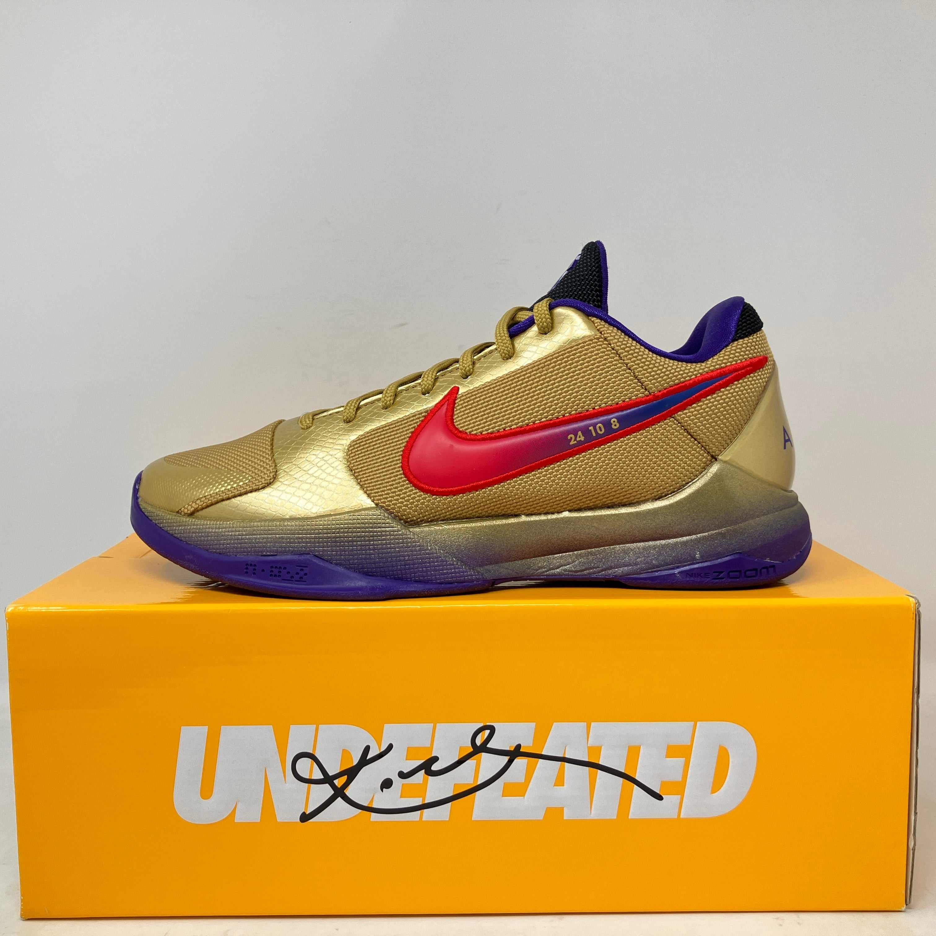 Nike Kobe 5 Protro basketball shoe in gold with red swoosh and purple accents on Undefeated branded box