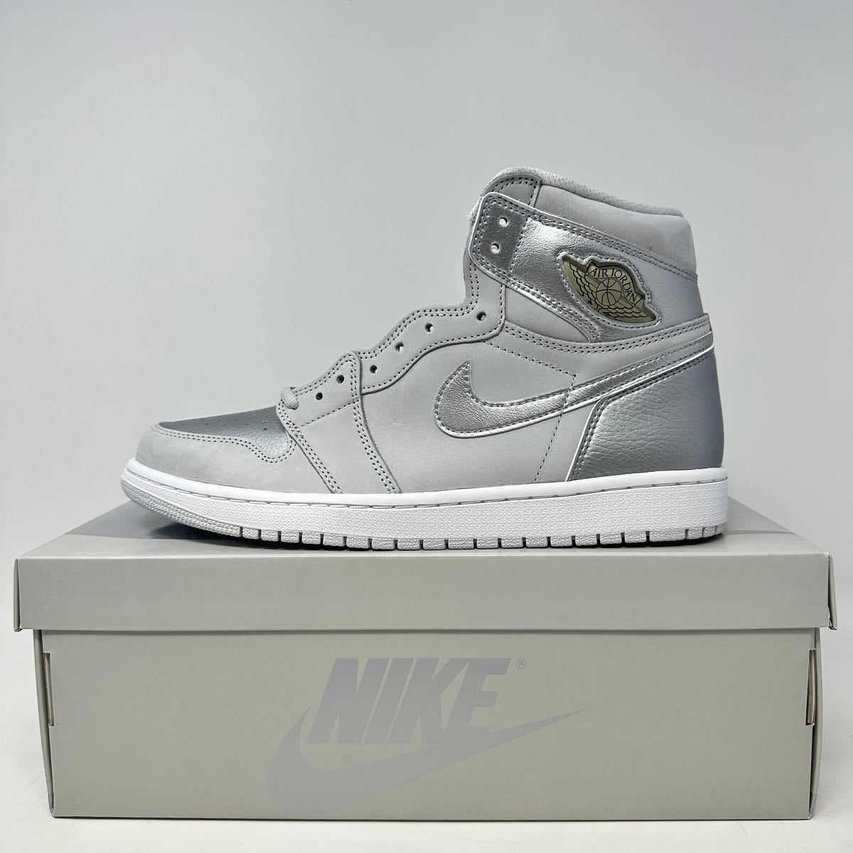 Jordan 1 Retro High sneaker in neutral grey leather with metallic accents, gold logo, and white sole on a grey Nike box.