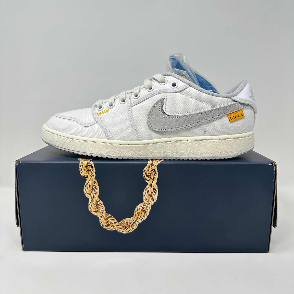 Jordan 1 Retro low-top sneaker in white canvas with a grey Swoosh, cream sole, yellow Union tag and exposed stitching.
