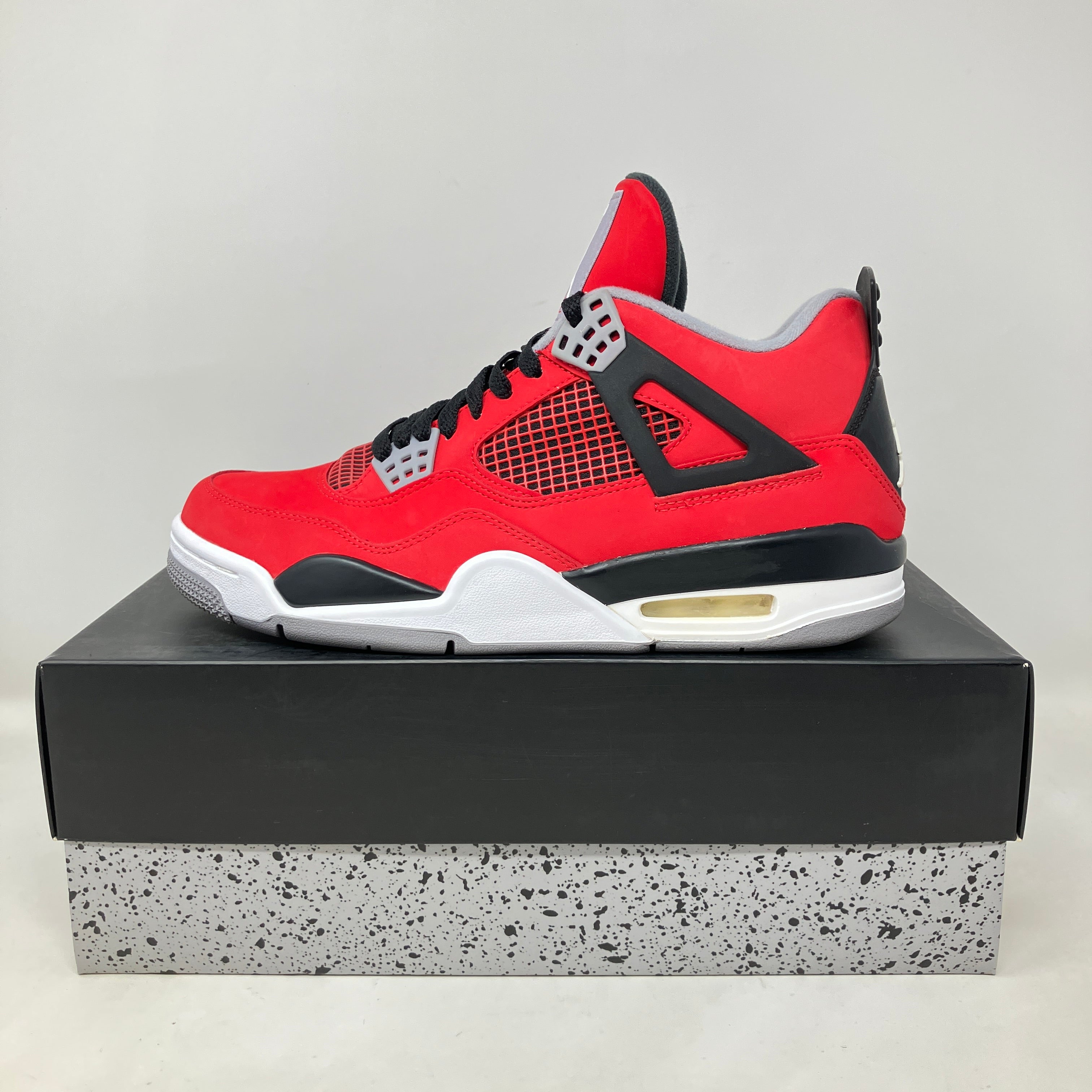 Red and black Jordan 4 Retro Toro Bravo sneaker on a black and speckled grey shoebox, pre-owned with all accessories included.