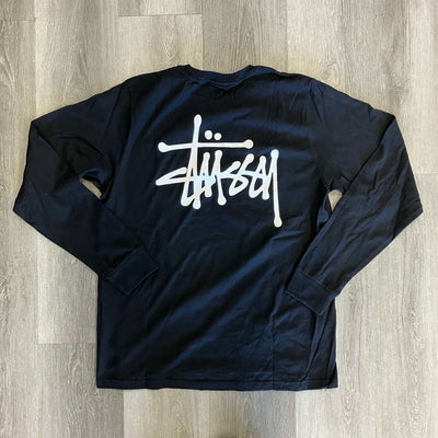 Black Stussy long sleeve tee with white logo on front, laid flat on wooden floor