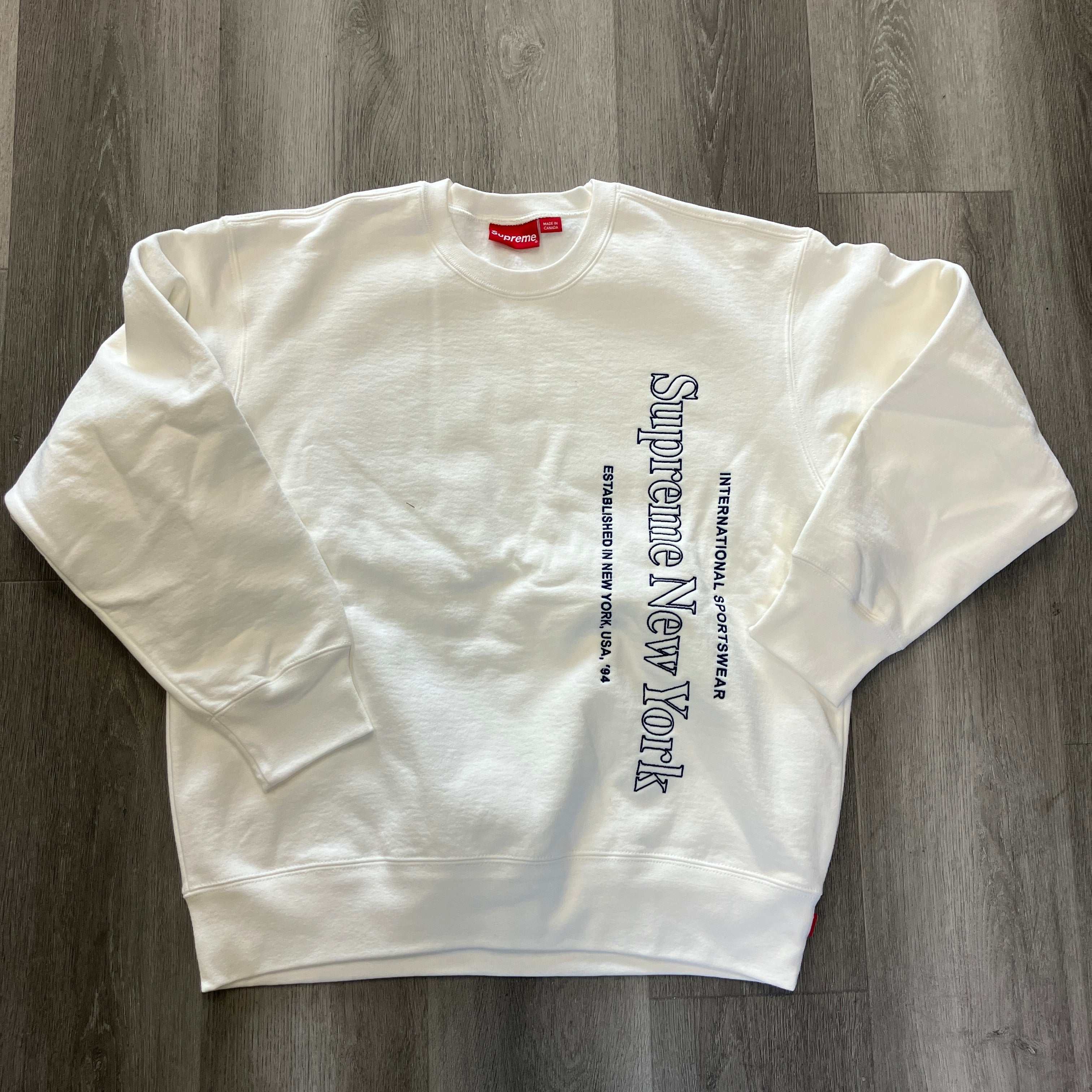 White Supreme Side Logo Crewneck sweatshirt with vertical navy text on front laid flat on wooden floor
