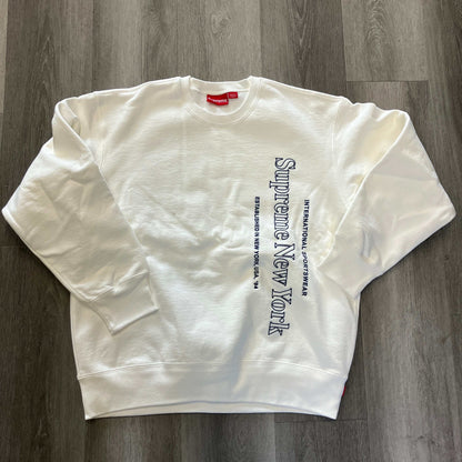 White Supreme Side Logo Crewneck sweatshirt with vertical navy text on front laid flat on wooden floor