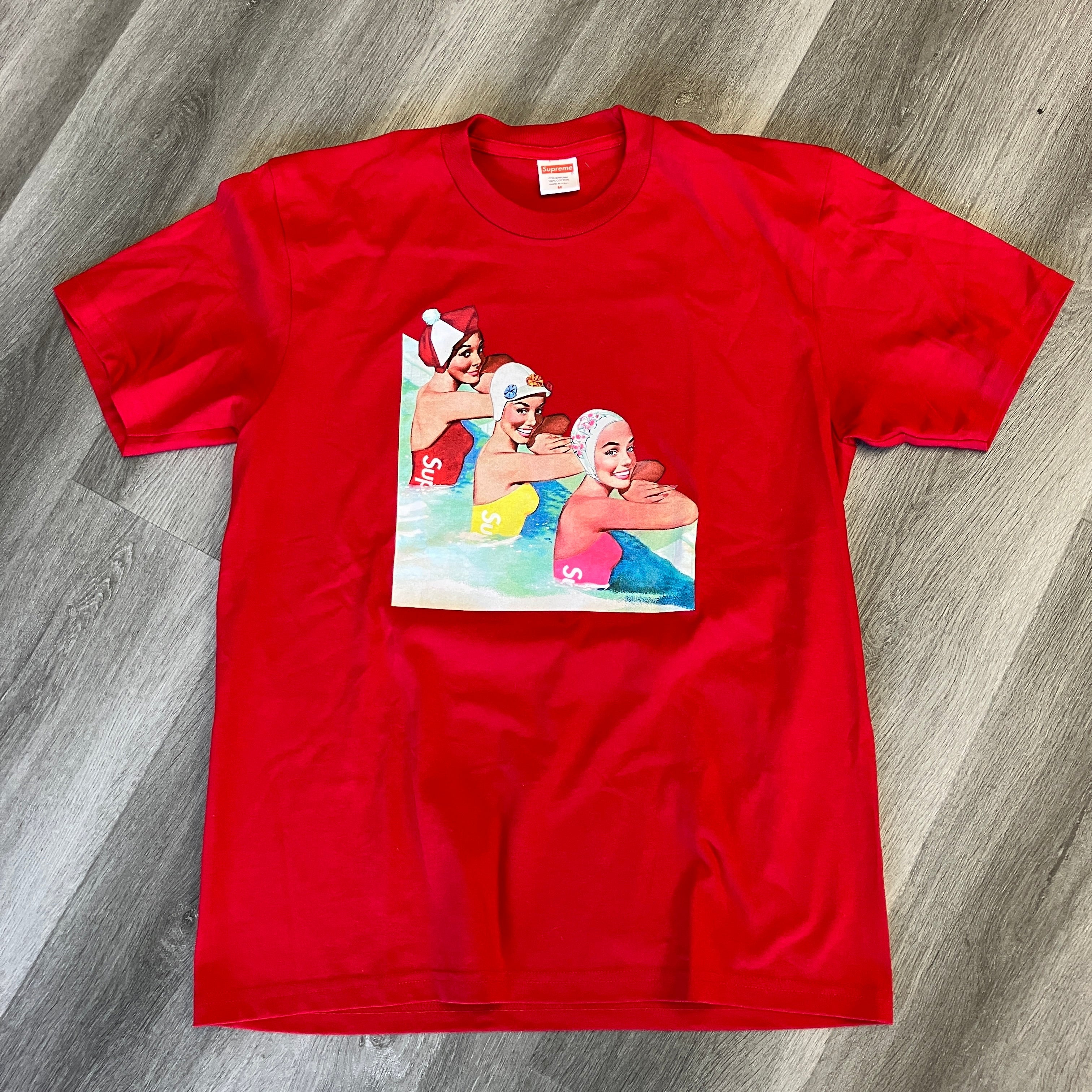 Red Supreme Swimmers Tee with graphic print of three swimmers in colorful swim caps, brand new, laid flat on wooden floor.