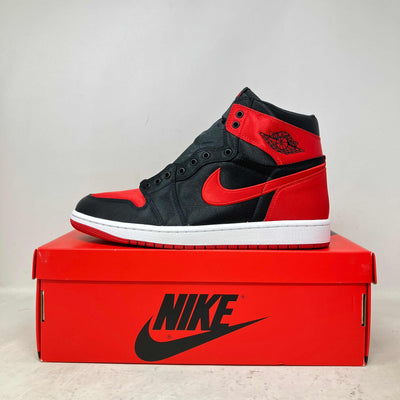 Jordan 1 Retro High OG sneaker in black and red satin material with a white sole and red Nike swoosh, displayed on a red Nike box.