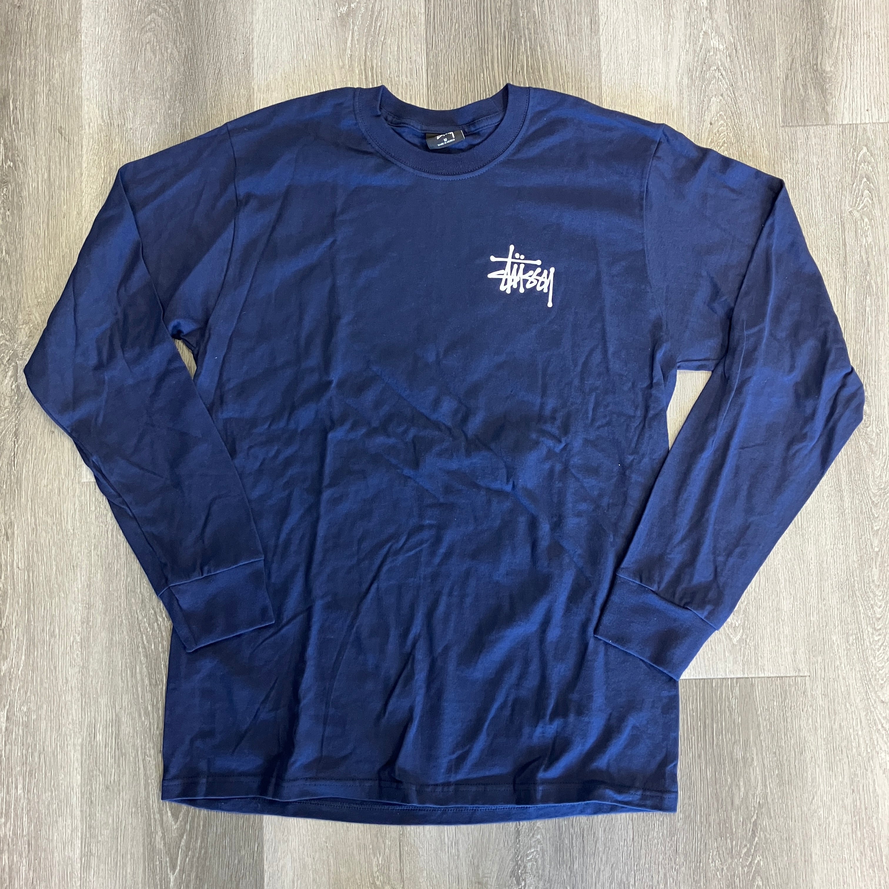Navy Stussy long sleeve tee with white logo on chest, brand new, laid flat on wooden floor