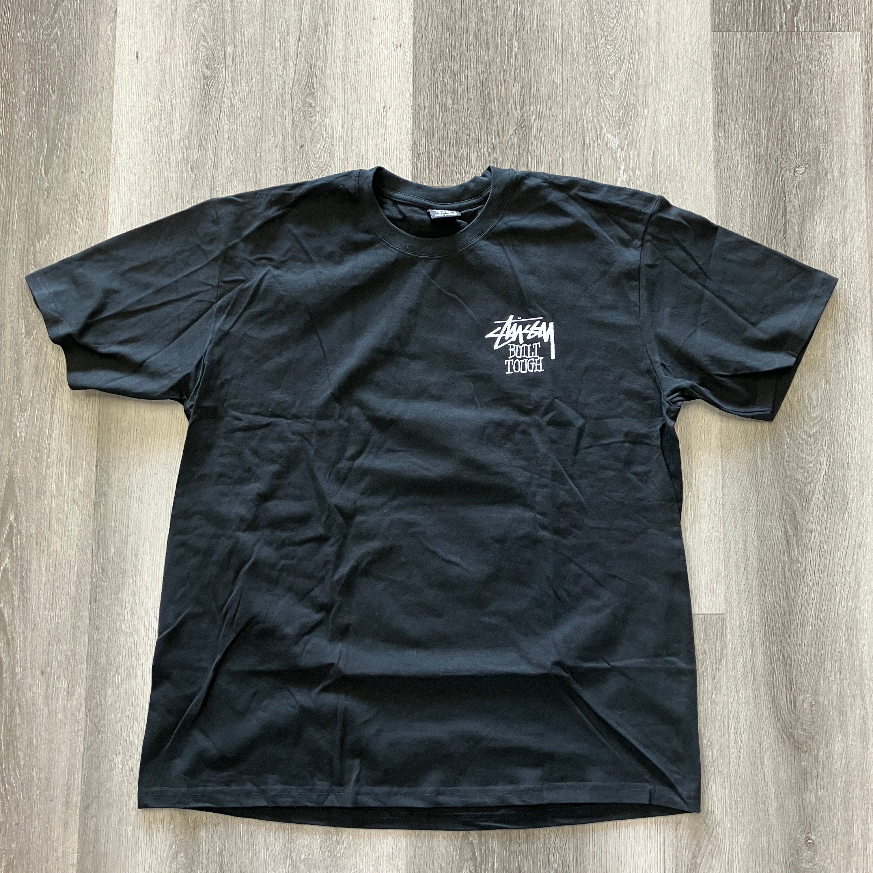 Black Stussy Built Tough Tee with white logo print, laid flat on wooden floor, brand new, 23.5in pit to pit, 32.5in length