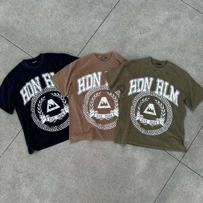 Three oversized Hidden Realm 'College' Tee shirts in black, brown, and olive green with white graphic print, laid flat on concrete.
